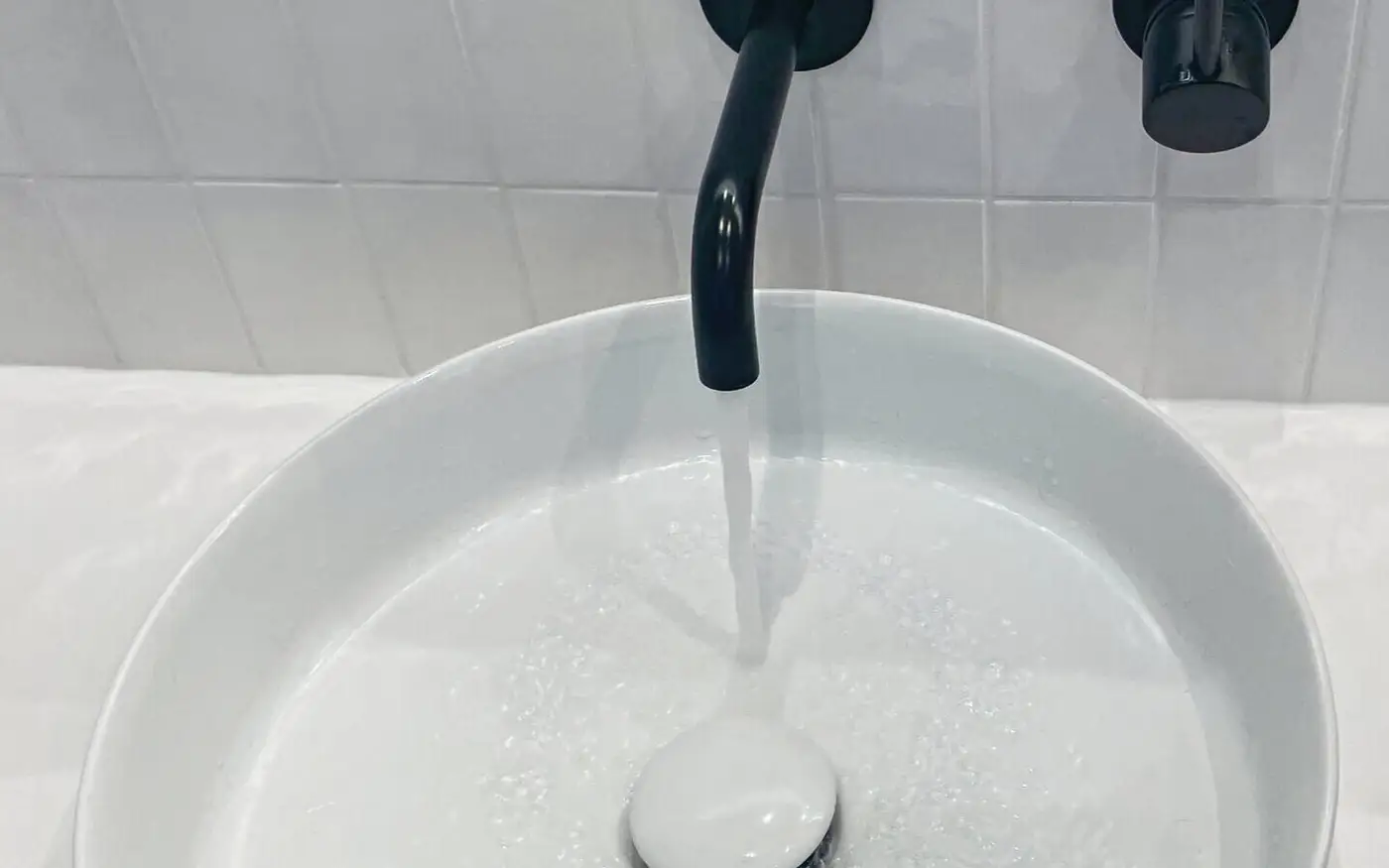 Round white vessel basin with a matte black wall-mounted tap running into the pop-up waste