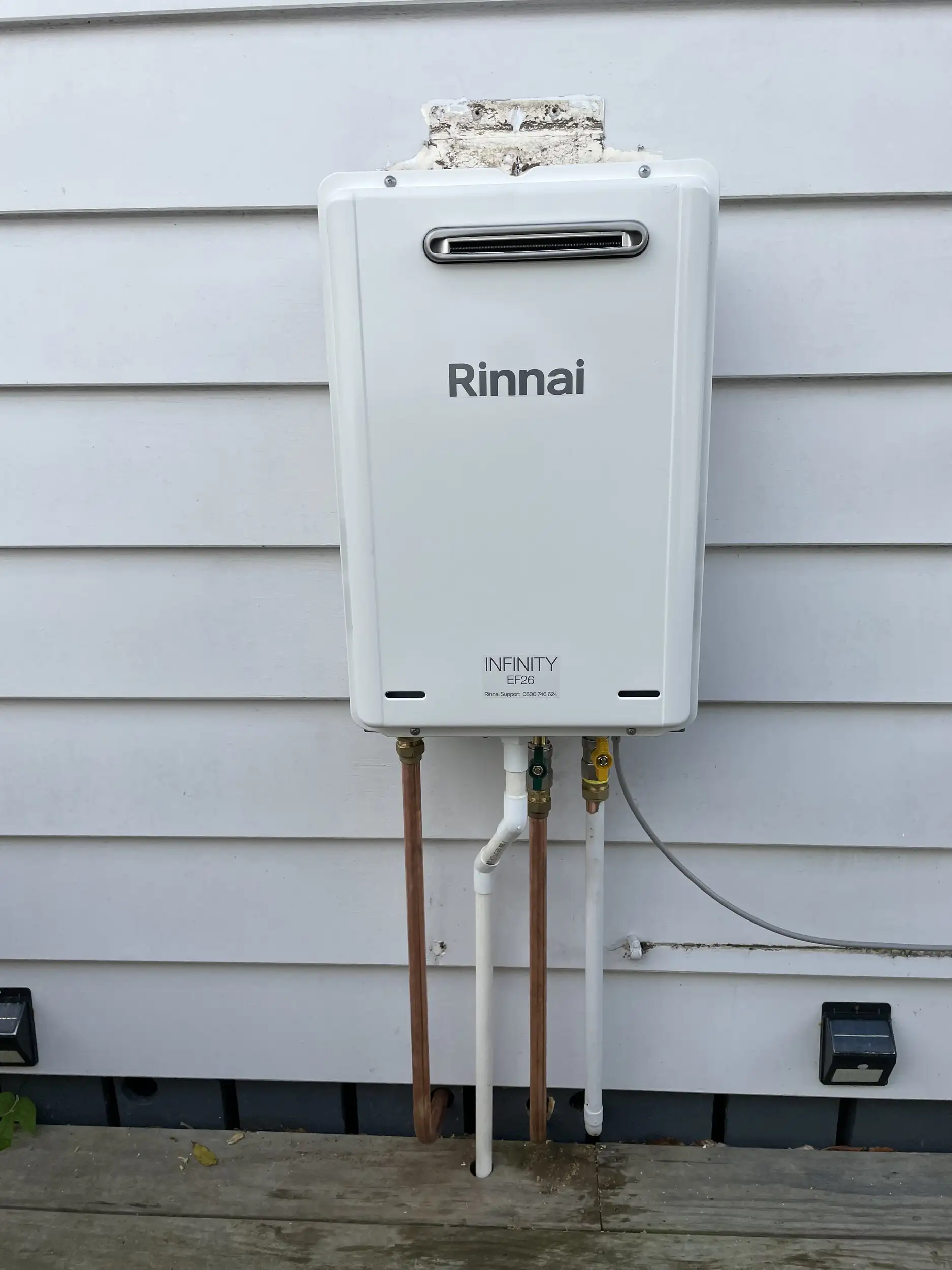 Rinnai INFINITY gas continuous flow water heater mounted on a weatherboard wall, with its pipework and outdoor power point below
